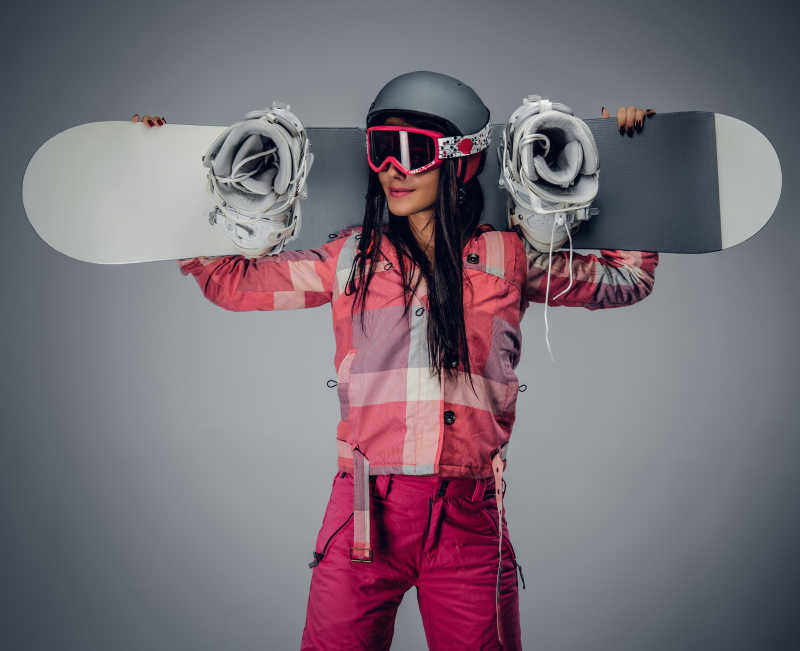 灰色背景下在肩上抱着滑雪板的女子