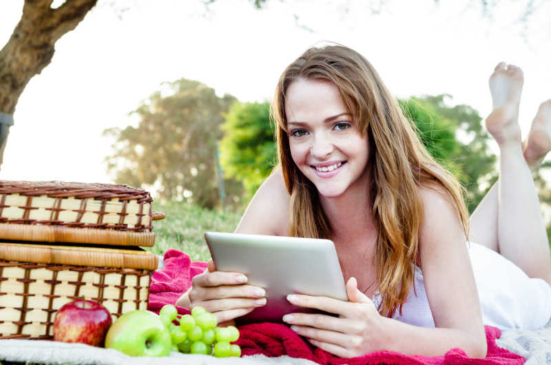 闲暇时光躺在公园的野餐毯上玩平板电脑的年轻女子