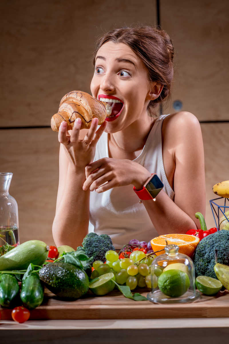 年轻女子在放满水果和蔬菜的桌子上吃甜的牛角包