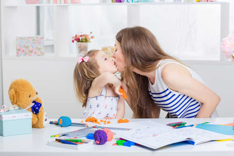 年轻的妈妈亲吻拿着橡皮泥的小女儿