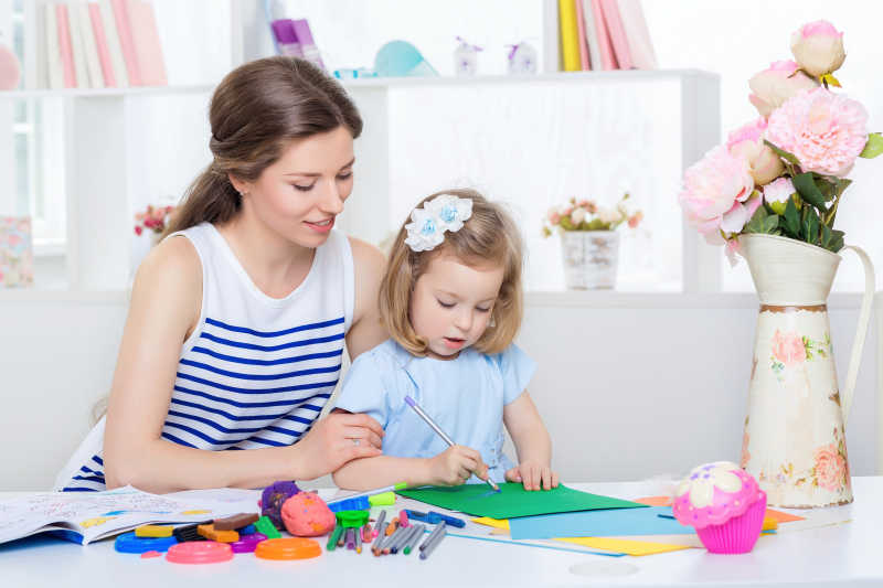 年轻美丽的妈妈微笑着看着她的女儿在彩纸上画画