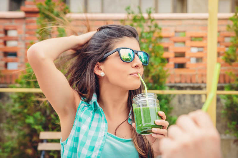 年轻漂亮的女人带着墨镜喝绿色蔬菜果汁