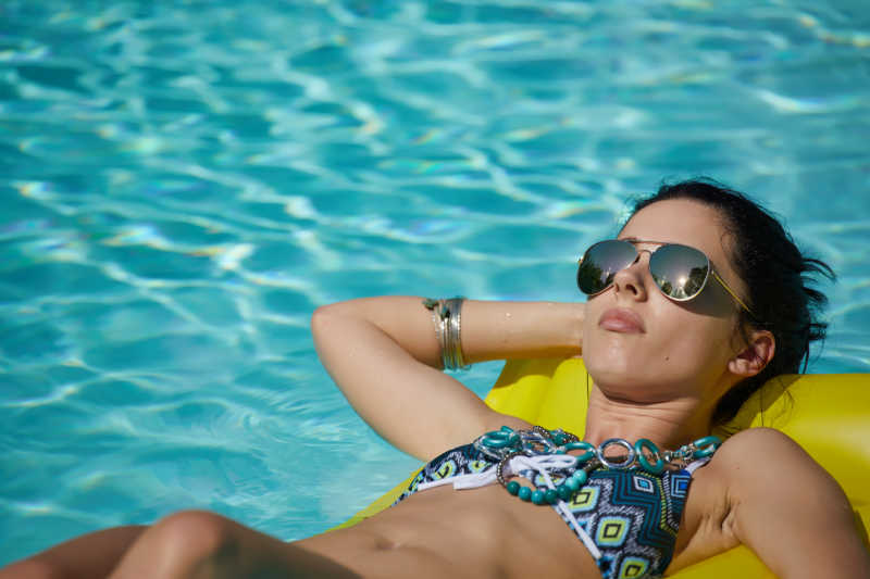 清澈的游泳池中空气床垫上戴着太阳镜躺着的比基尼女郎