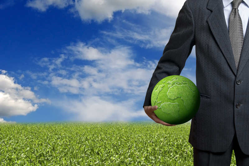 蓝天白云背景下站在绿植中拿着绿色地球模型的黑衣商人
