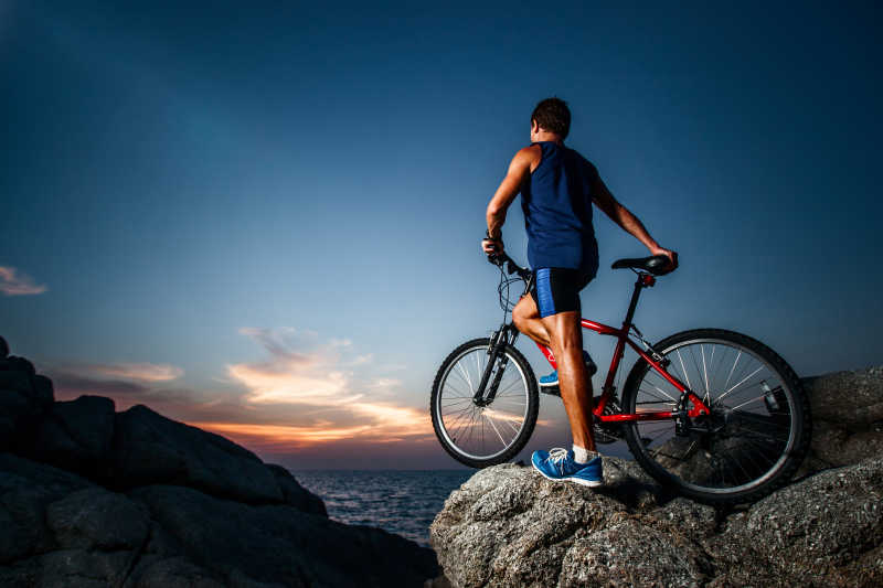 日落时分骑着自行车站在岩石上的年轻男子