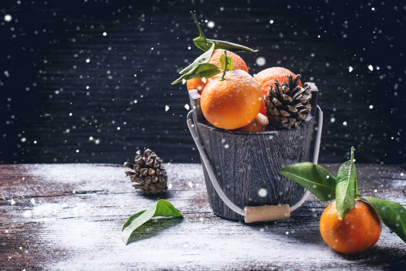 雪天黑色背景下的木桶里的橘子和松果