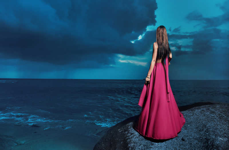 穿着礼服的的少女站在岩石上看着平静的海面