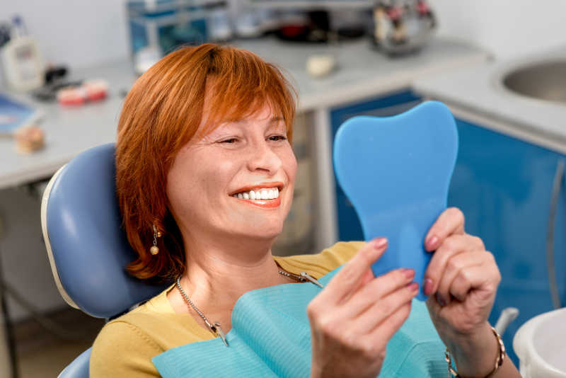 带微笑的老妇人坐在牙科诊所看着镜子