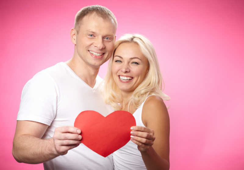 粉色背景下幸福年轻夫妻手拿红色的纸心的画像