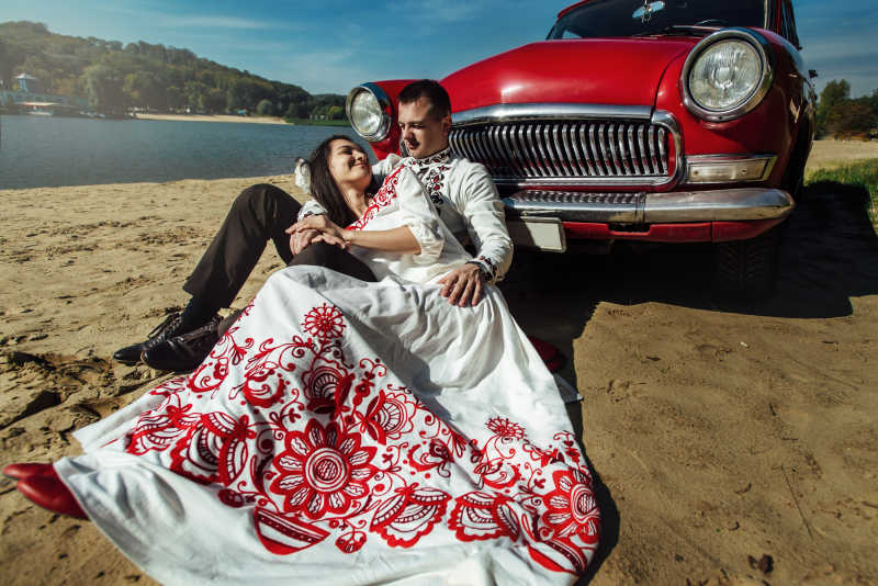 沙滩上坐在红色汽车旁边的新郎和新娘