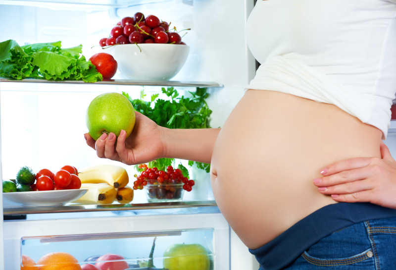 站在冰箱边吃水果蔬菜的孕妇