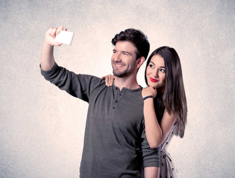 在一个空的清灰墙背景概念前的一对年轻夫妇拿着手机自拍