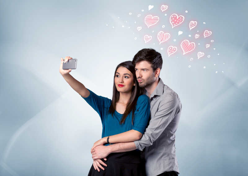 恋爱中的年轻夫妇和绘制红心在一个空的清灰墙背景概念前的帅哥的手机