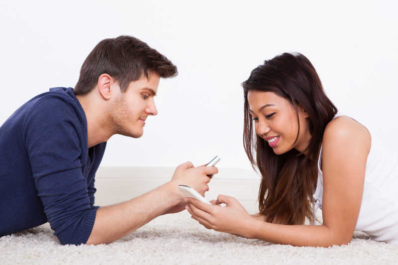 年轻夫妇相对的趴在白色地毯上玩手机