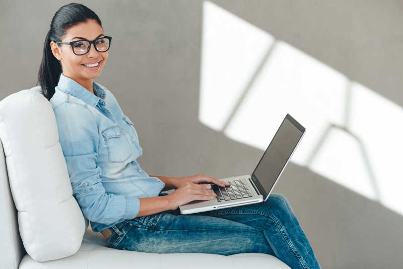 坐在沙发上的美丽的年轻女子戴着眼镜使用笔记本电脑