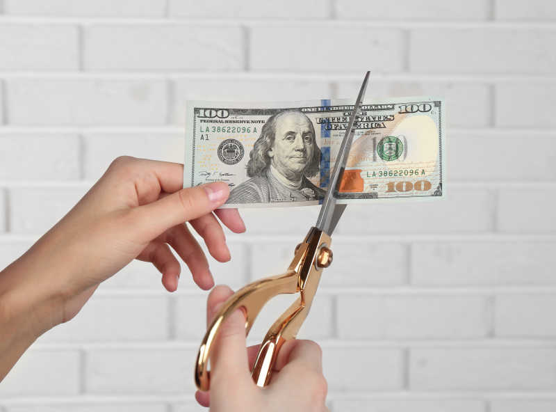 双手用剪刀在白色砖墙的背景下切钱