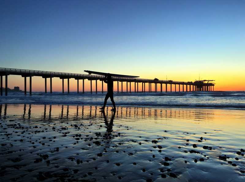 日落时分搁浅海滩上拿着冲浪板走着的年轻人