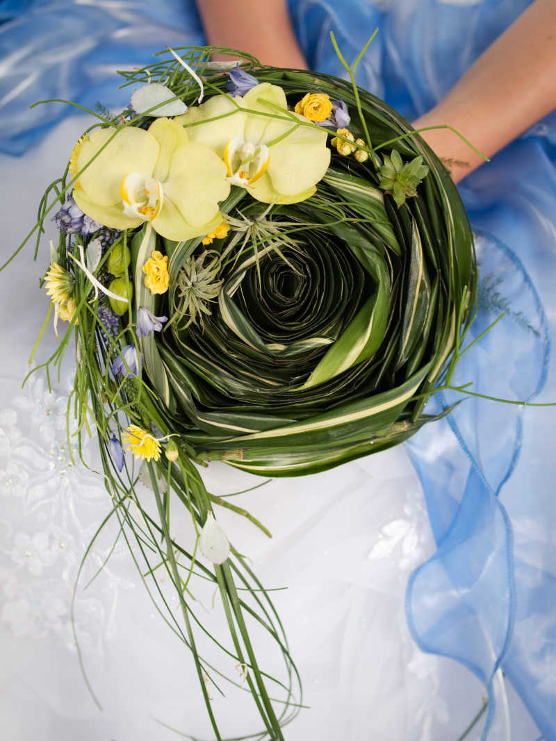 穿着蓝色和白色婚纱的新娘手中的黄色兰花和绿叶的婚礼花束