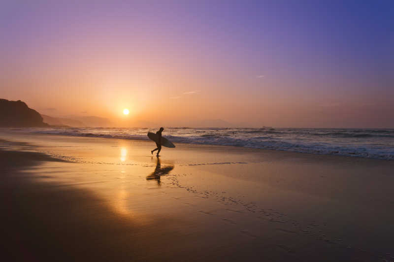 日落时拿着冲浪板走在沙滩上人