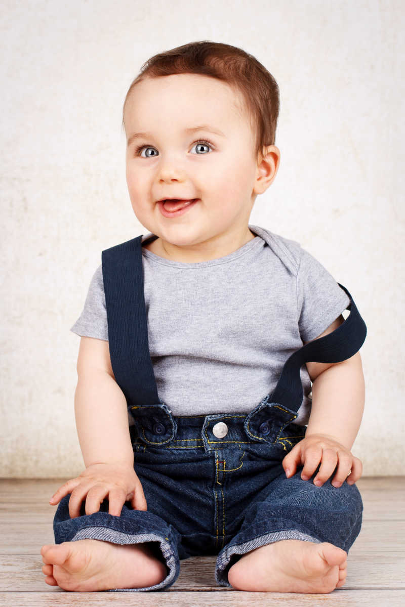 白色背景下坐在地板上穿着牛仔背带裤微笑的宝宝