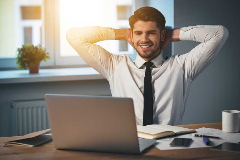 英俊活泼的年轻人坐在办公室双手紧握在头上微笑着