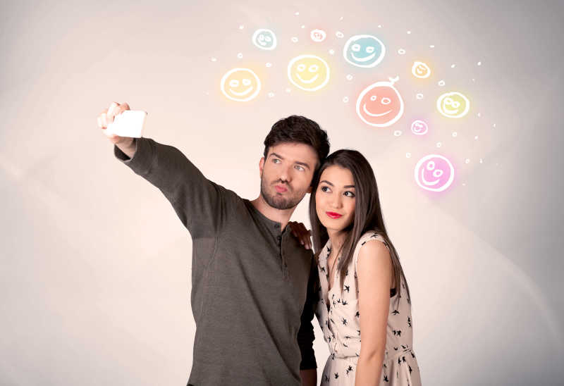 在多姿多彩的幸福笑脸图背景下拿着手机自拍的年轻夫妇