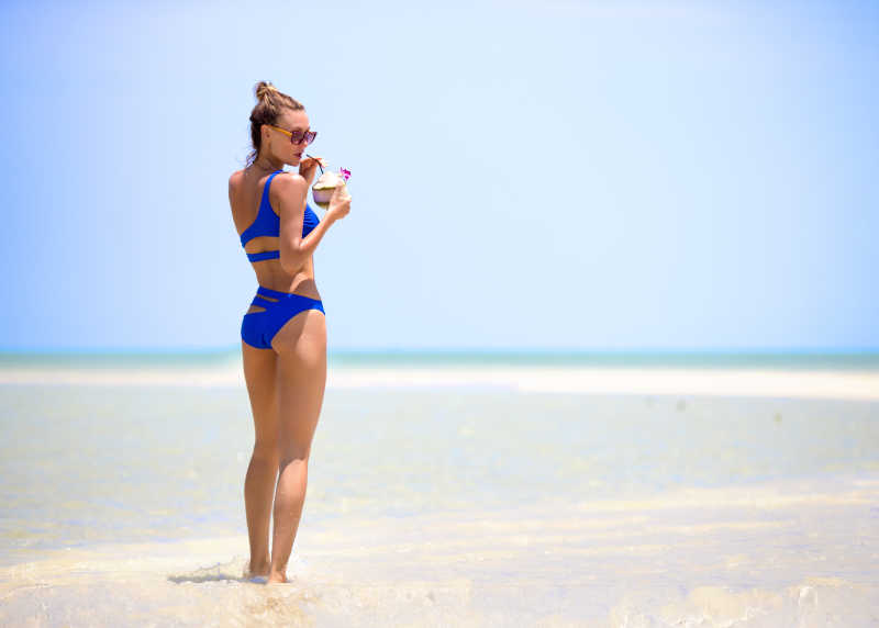 蓝天下金色沙滩上穿着蓝色比基尼享受美食的美女