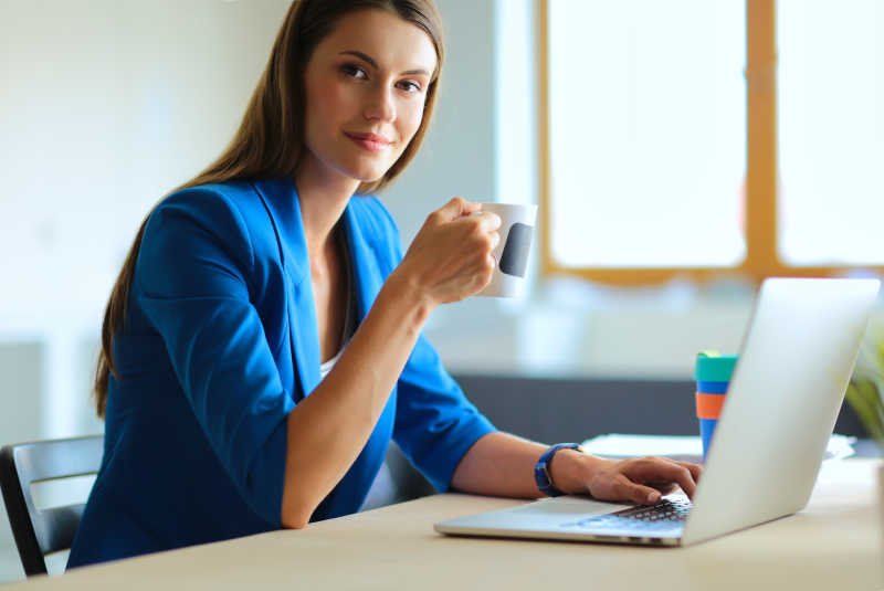 微笑着的女商人坐在办公桌旁捧着一杯咖啡