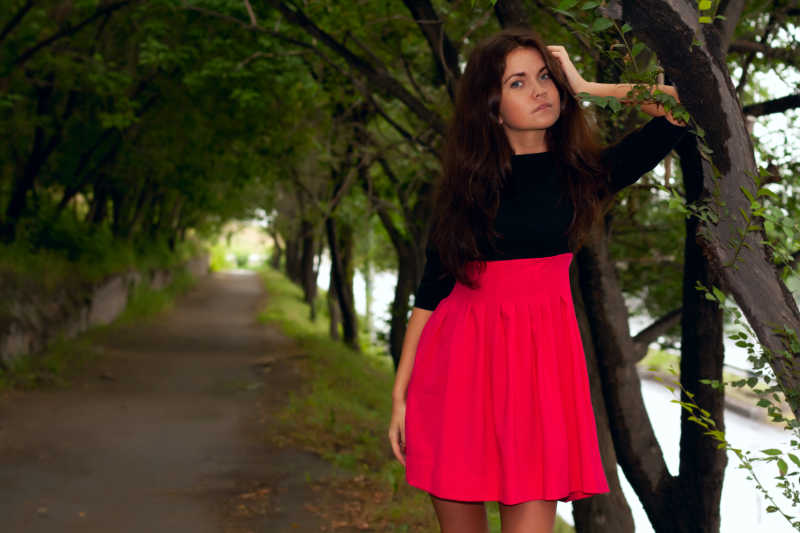 美丽的调情女人穿着红色连衣裙走在公园的小巷