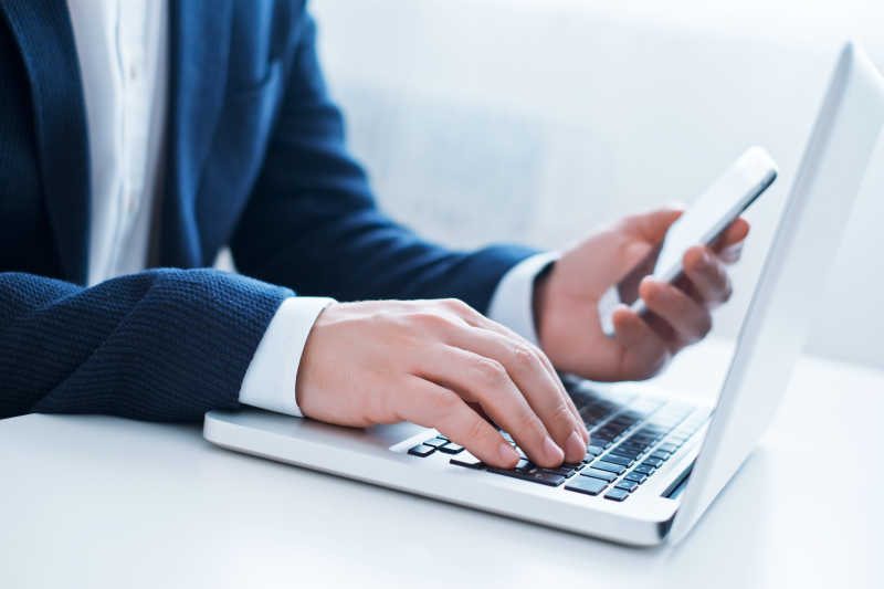 白色桌上的男人一边使用智能手机一边在笔记本电脑上打字