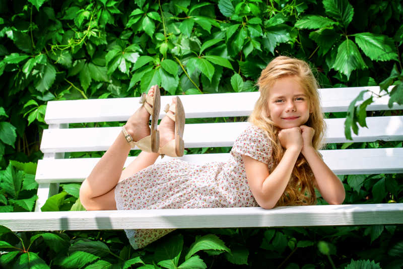 绿色树叶背景下趴在白色长椅上的金发小女孩