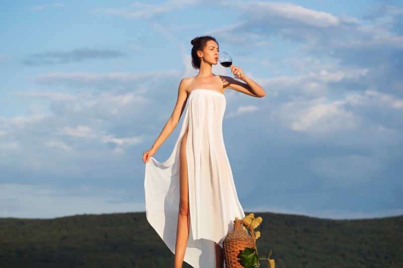 蓝天白云下户外站在柳条瓶后面喝着葡萄酒的性感美女