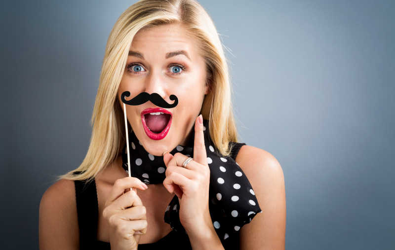 灰色背景下拿胡子样式纸党棍拍照的搞怪金发美女