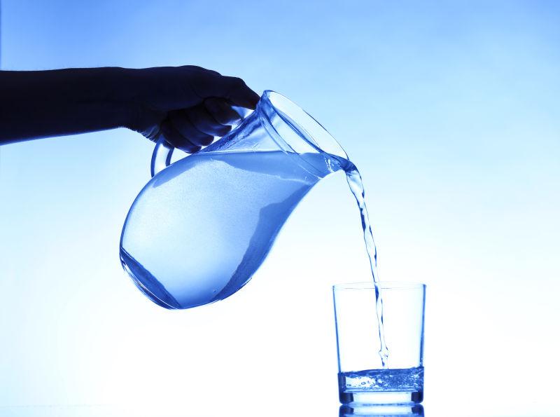 在深蓝色背景下将水罐中的水倒入玻璃杯中