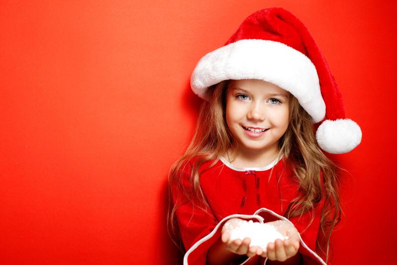 红色背景下戴着圣诞帽的小女孩手里捧着雪