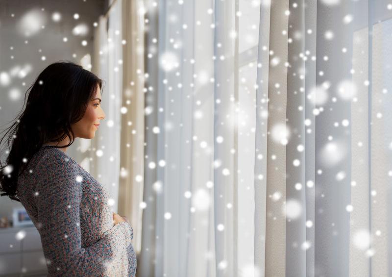 快乐的孕妇怀着大肚子望着窗外的雪