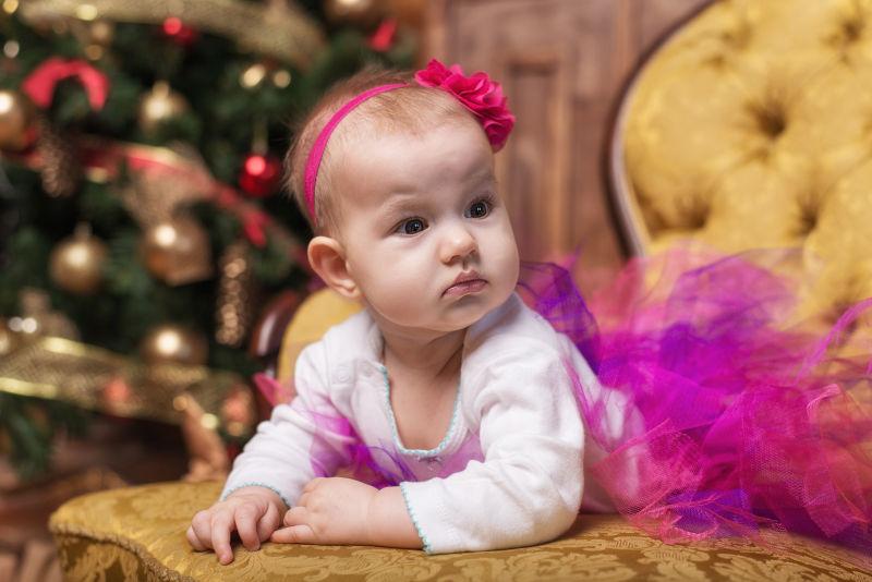 可爱的小女孩穿着粉红色的裙子和红色的头巾躺在圣诞树前的沙发上