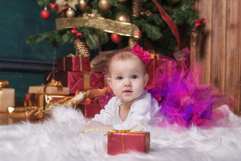可爱的小女孩穿着粉红色的裙子和红色的头巾爬在白色地毯上的圣诞树下