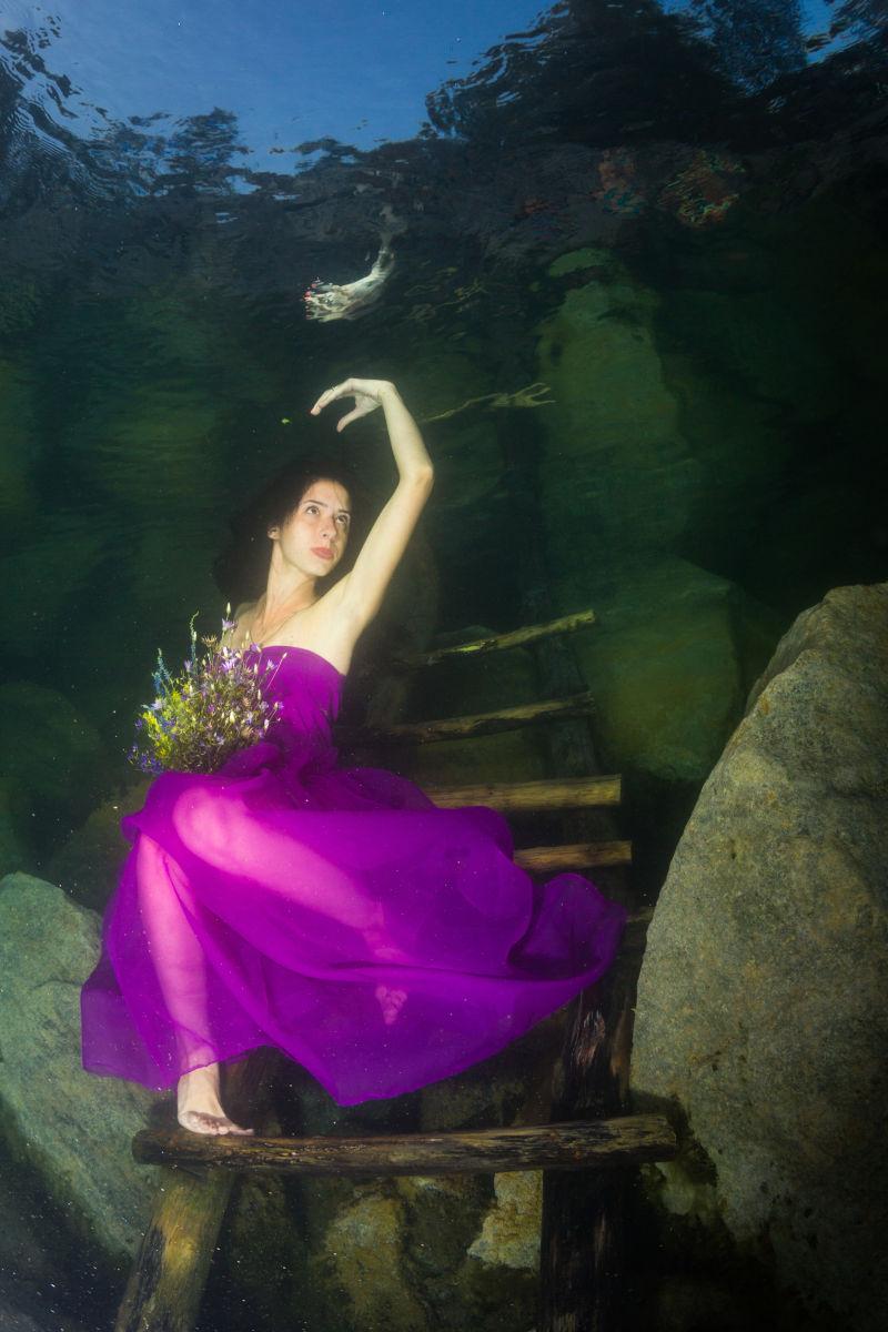 穿着紫色裙子拿着一束花的年轻优雅的女人在水下楼梯上