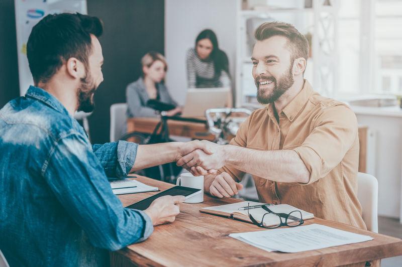 两个自信的年轻人一边坐在办公桌前一边握手