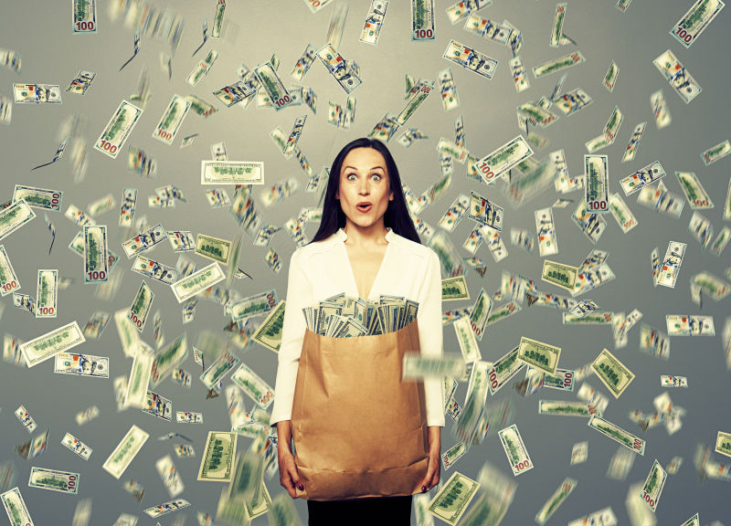 惊讶的年轻女商人在美元雨下拿纸袋和钱