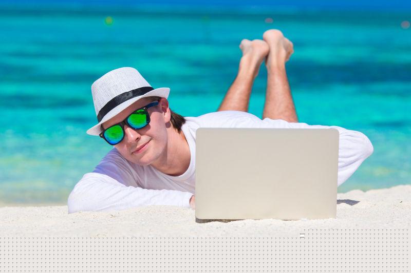 阳光下趴在沙滩上使用笔记本电脑的男人