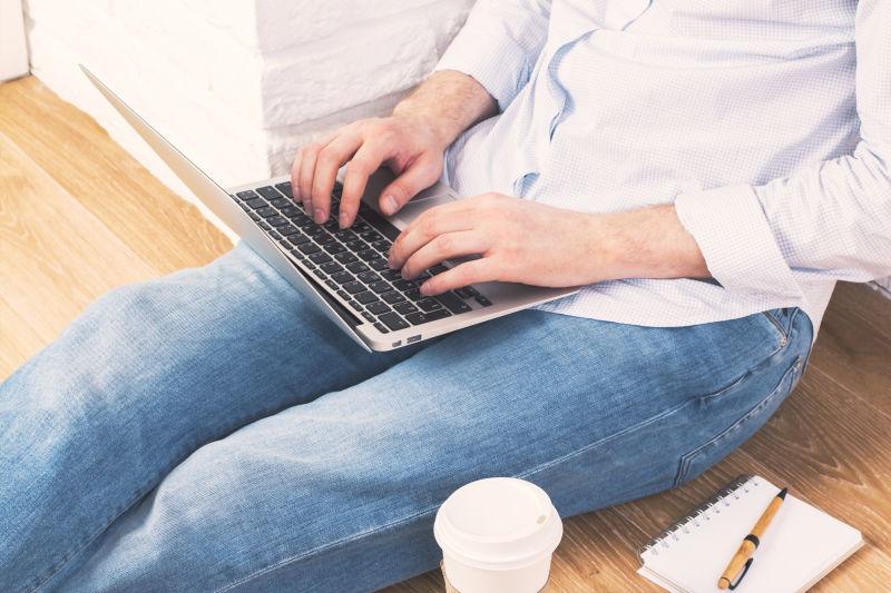 在腿上使用笔记本电脑键盘打字的坐在木地板上的男人
