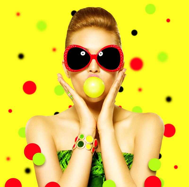 有彩色泡泡糖背景的少女戴太阳镜吃泡泡糖