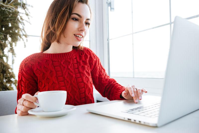 穿红毛衣的年轻美女一边喝咖啡一边使用笔记本电脑