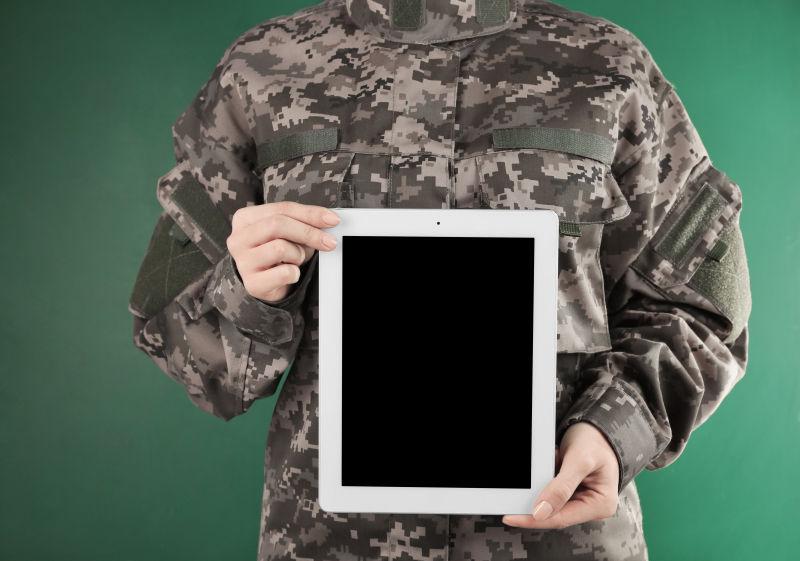 绿色背景墙前拿着平板电脑的迷彩装男人