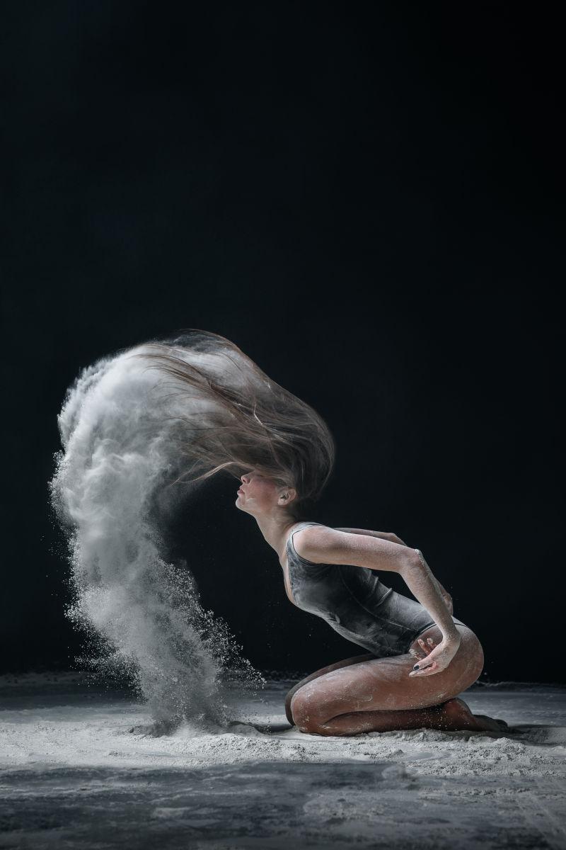 黑色背景下跪坐在地上使用头发甩出面粉的舞者