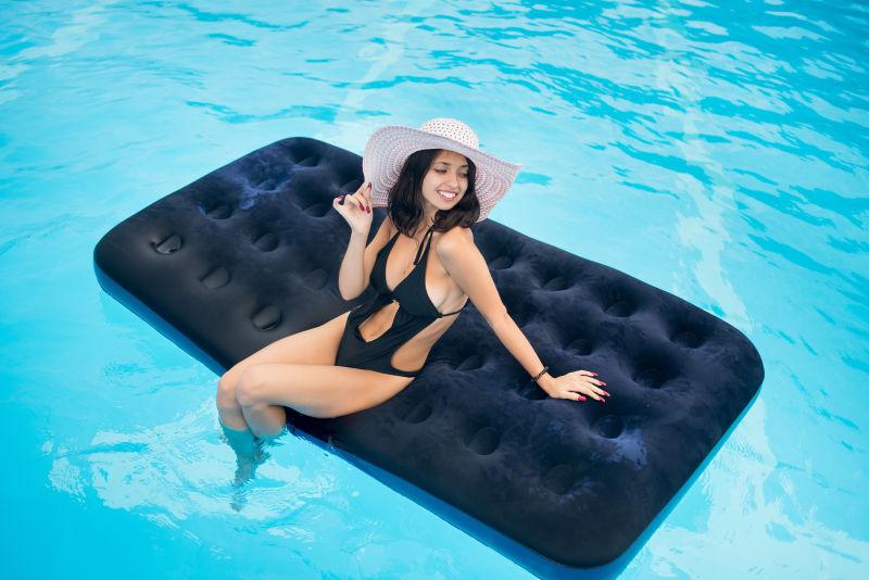 穿着黑色比基尼的女人戴着帽子坐在泳池里的床垫上