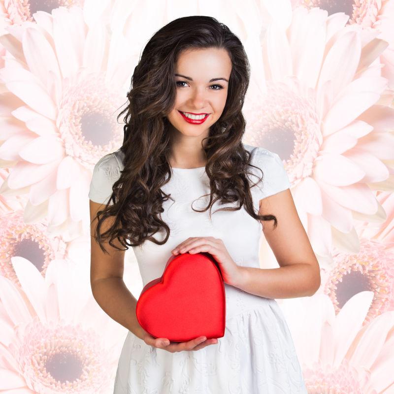 粉色菊花花朵背景下穿着白色衣裙抱着红色心形盒子的美女