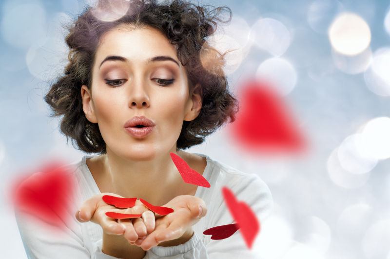 散景背景上年轻美女吹着手心的红色心形纸片
